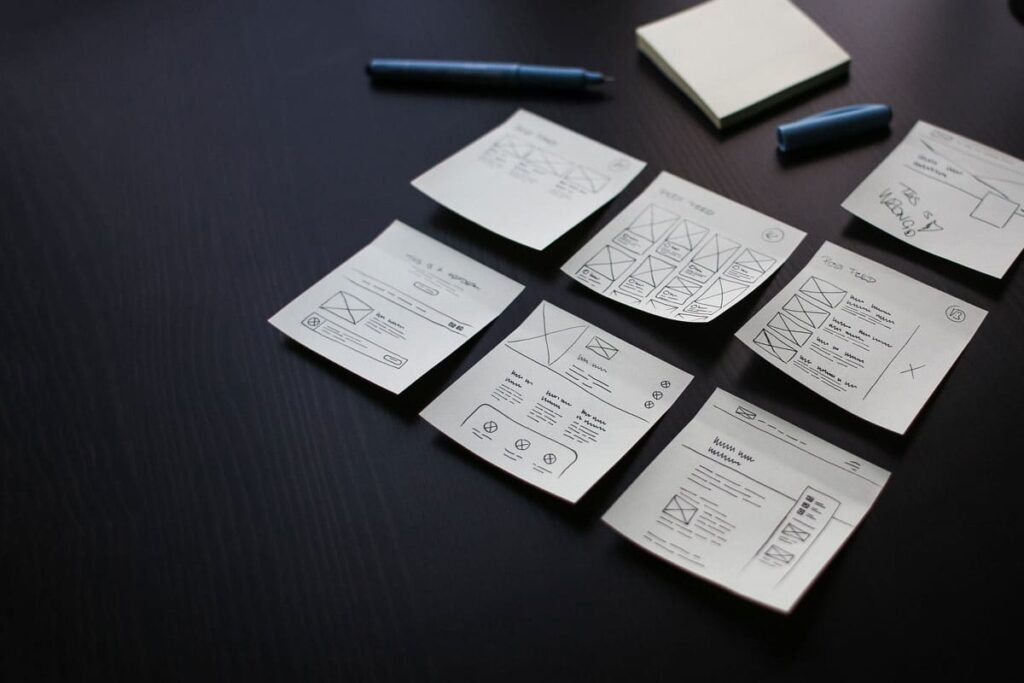Sticky notes sketched with website wireframes arranged on a dark desk, with a pen and notepad nearby