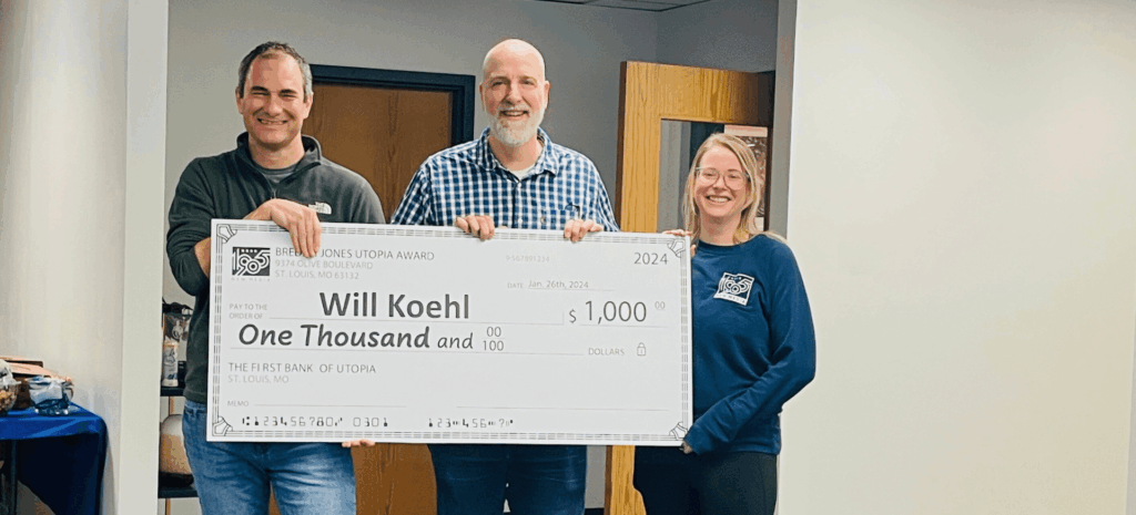 Three colleagues holding an oversized 1905 New Media check made out to Will Koehl for 1,000 dollars, standing in an office hallway.