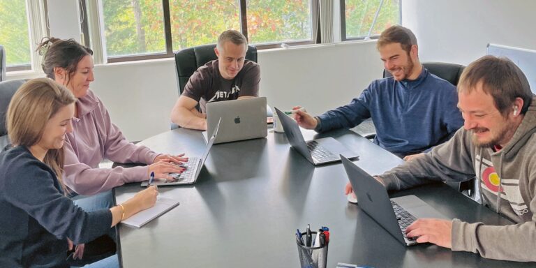 1905 New Media team members collaborating around a conference table with laptops and notebooks in a bright office with large windows.