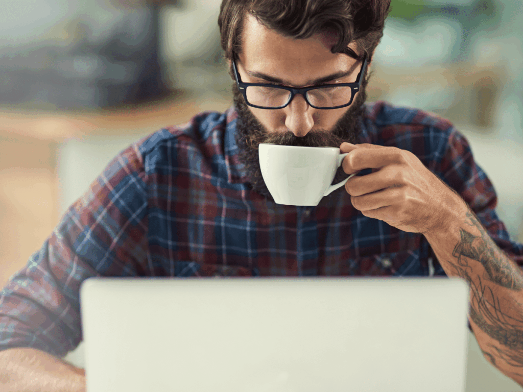 Person in a plaid shirt working at a laptop and drinking from a white mug, with a tattooed forearm visible.