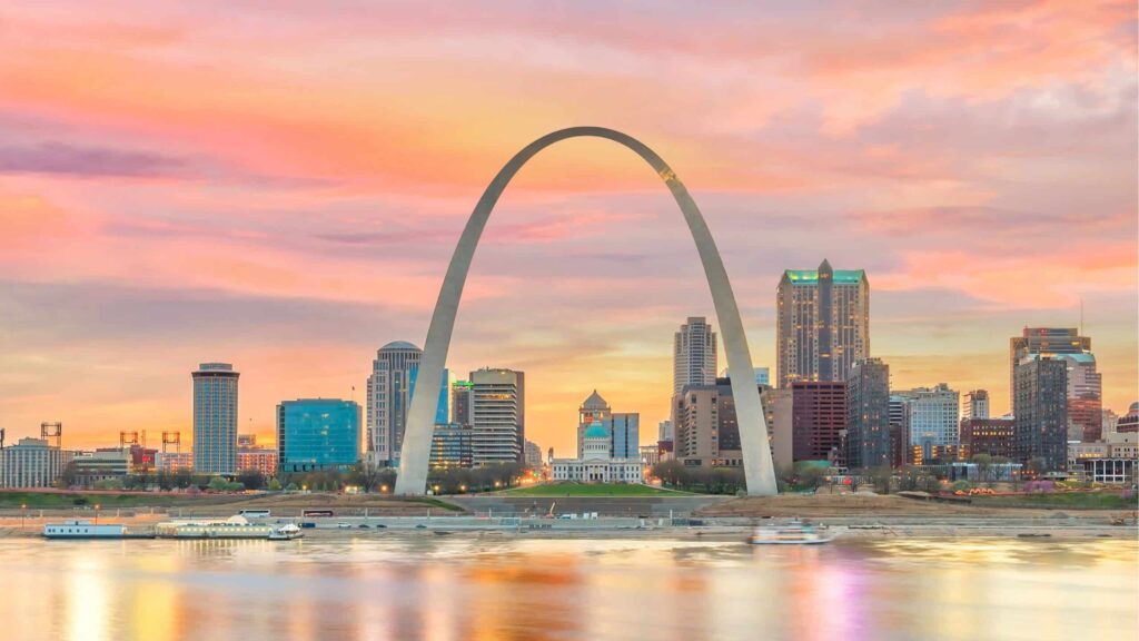 St. Louis skyline at sunset with the Gateway Arch centered in front of colorful pink and orange clouds reflecting on the Mississippi River.