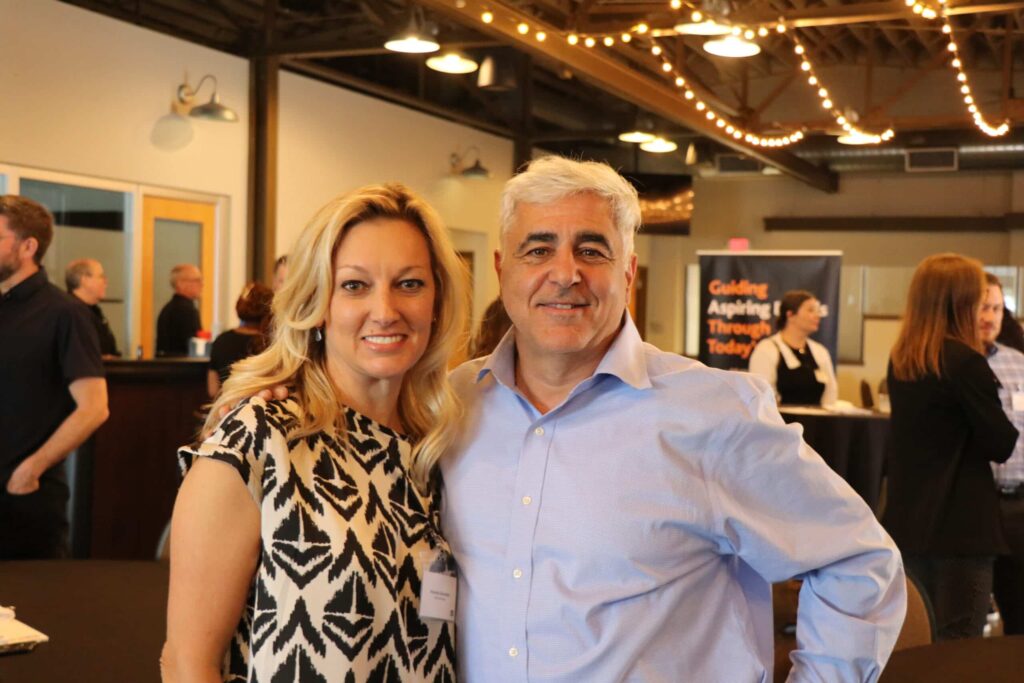 Two Marketing Disrupted attendees stand close together and smile for the camera at a networking reception, with other guests and warm event lighting softly blurred in the background.