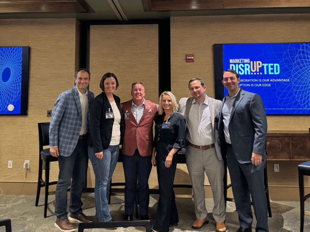 Group photo of panelists and hosts standing together at a Marketing Disrupted generational business leaders event in St. Louis, with conference branding displayed on screens in the background.
