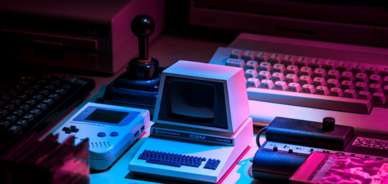 Miniature Commodore CBM computer replica next to a classic Nintendo Game Boy and a retro joystick under vibrant neon lighting.
